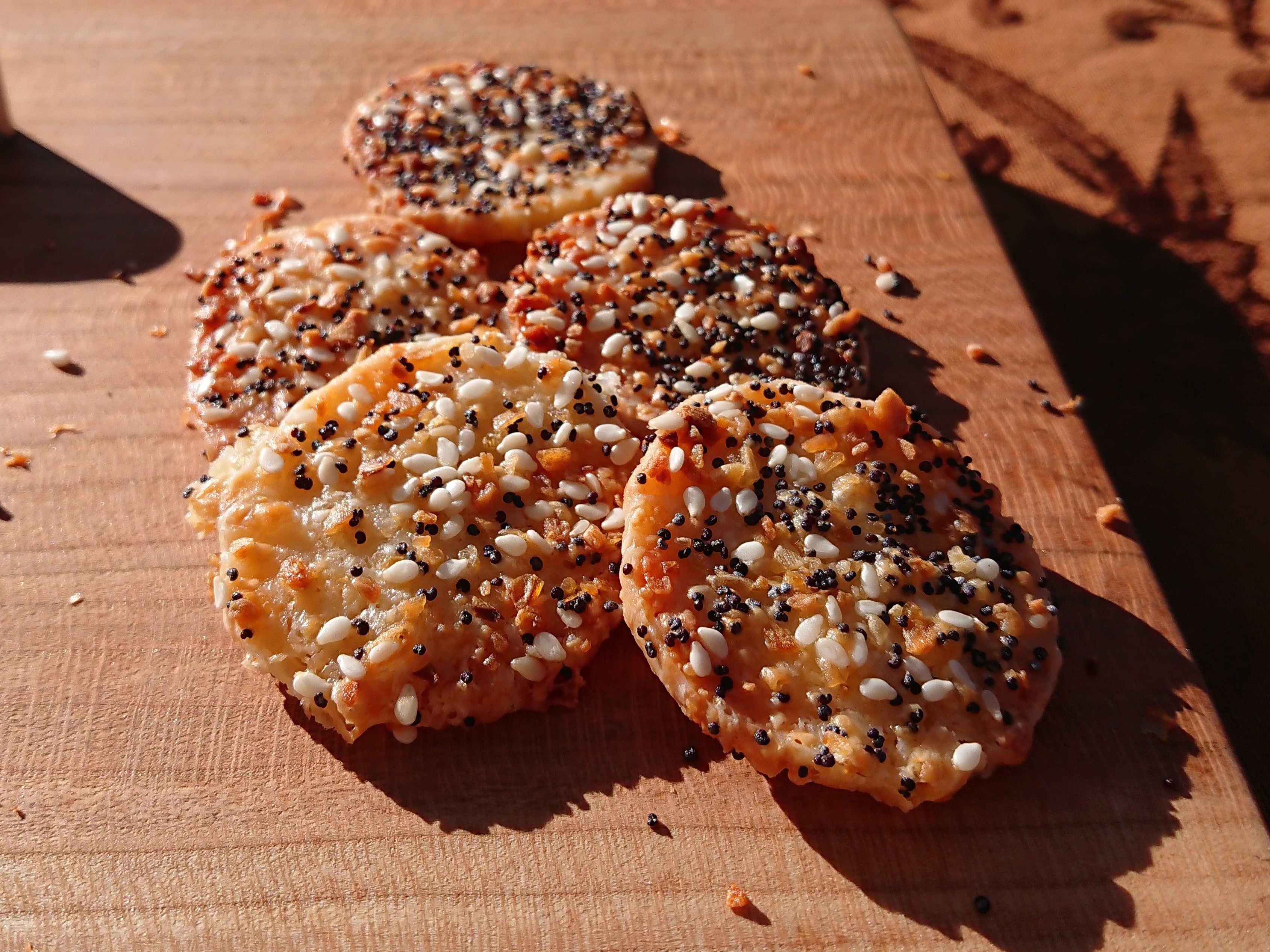 Five round crackers topped with mixed seeds and spices, arranged on a wooden board outdoors on a patterned tablecloth with a blurred green field in the background.