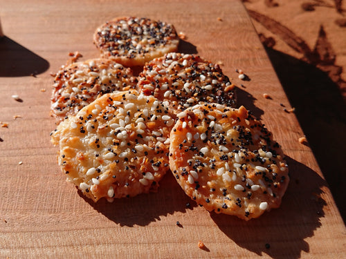 Five round crackers topped with mixed seeds and spices, arranged on a wooden board outdoors on a patterned tablecloth with a blurred green field in the background.