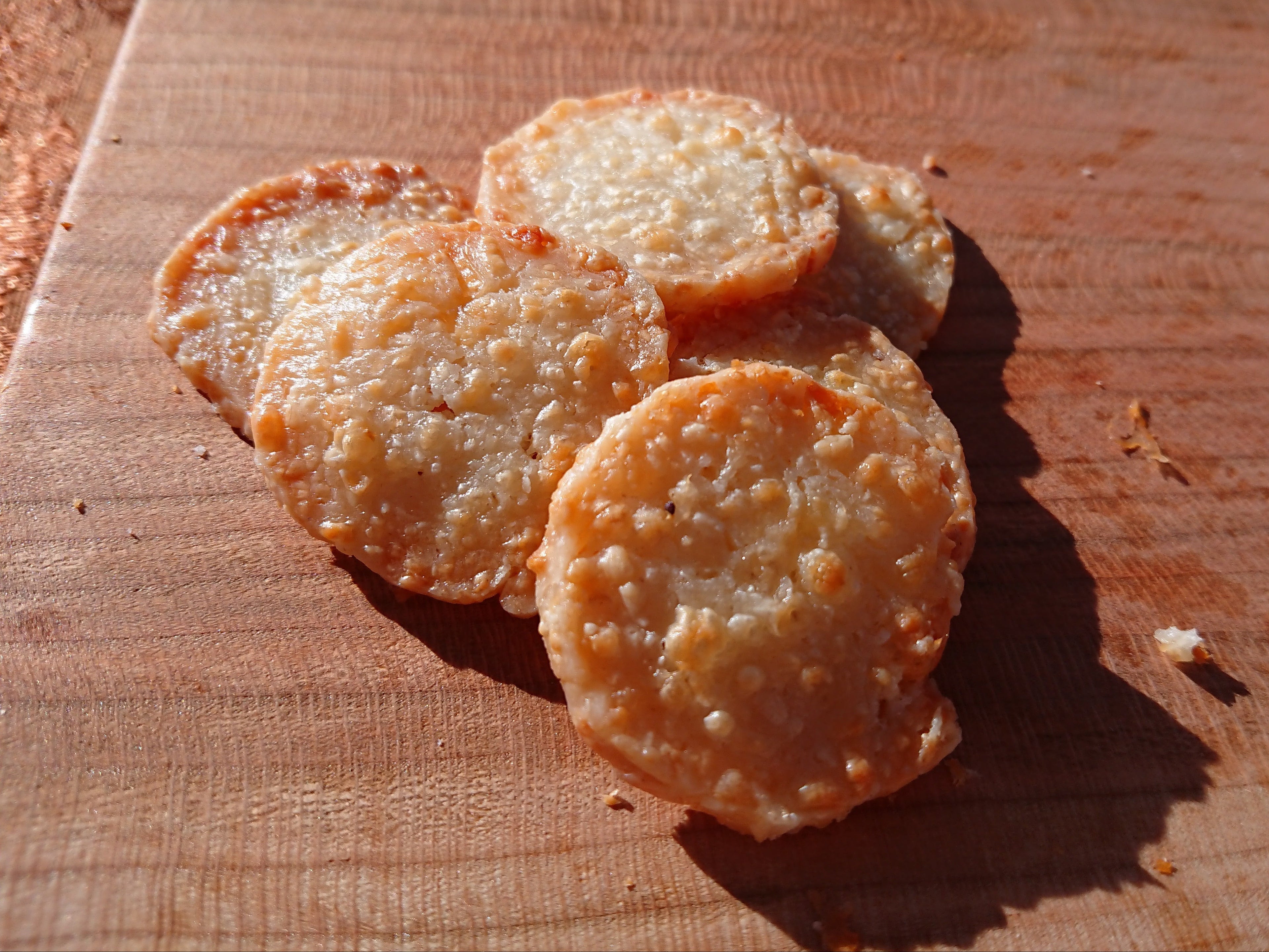 Six golden-brown, round, crispy baked crackers arranged on a wooden cutting board under direct sunlight.