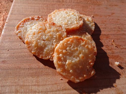Six golden-brown, round, crispy baked crackers arranged on a wooden cutting board under direct sunlight.